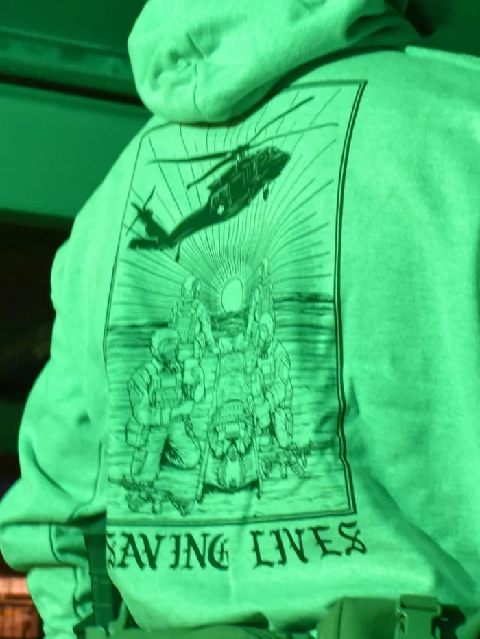 Military-style hoodie featuring the Saving Lives design: a group of soldiers carrying a stretcher while a helicopter prepares to land, symbolizing courage under pressure.